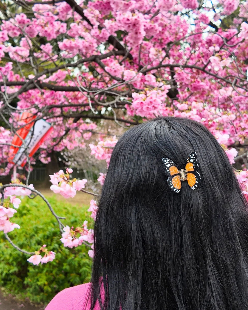 Orange-and-black butterfly claw hair clip securing long black hair, with pink flowering branches in the background.
