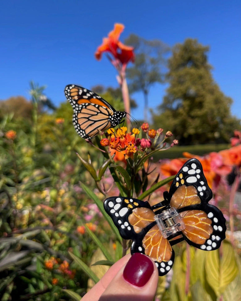 Orange-and-black butterfly claw hair clip held in front of orange flowers, with a monarch butterfly perched on the flowers behind it.