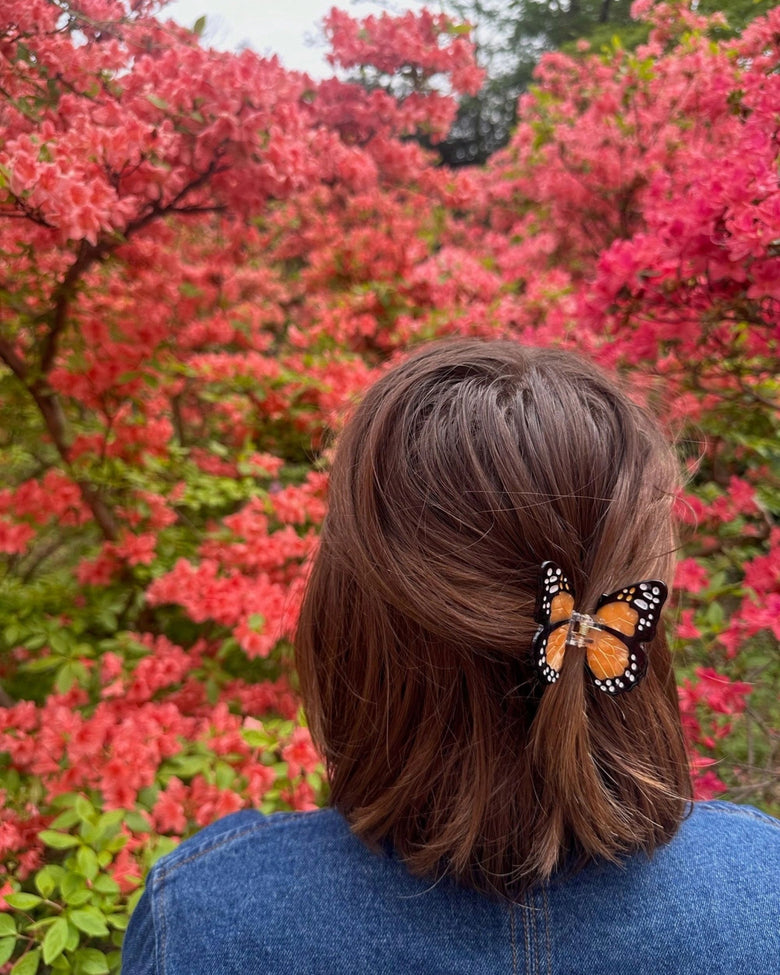 Orange-and-black butterfly claw hair clip securing a section of brown hair, with red flowering shrubs behind.