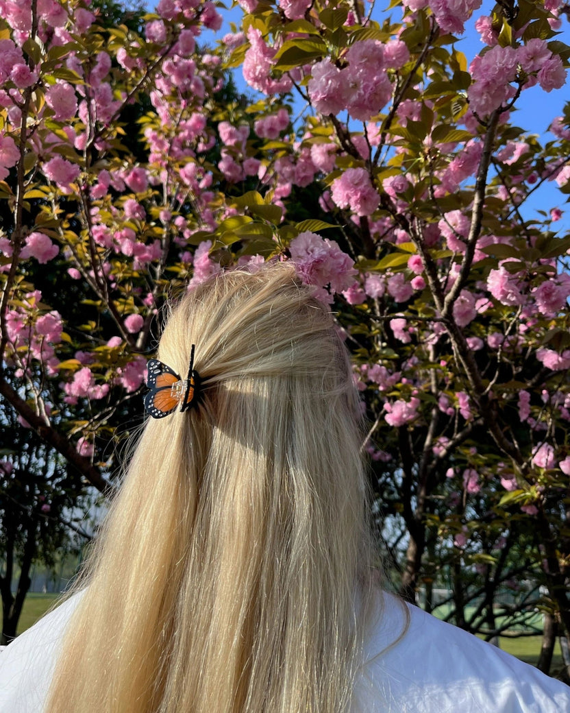 Orange-and-black butterfly claw hair clip securing a half-up section of long blonde hair, with pink flowering branches behind.