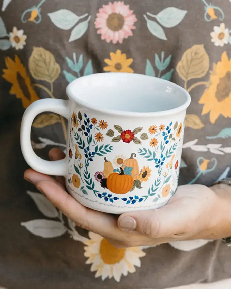 White ceramic mug with a pumpkin, flowers, leaves, and berries design, held in two hands.