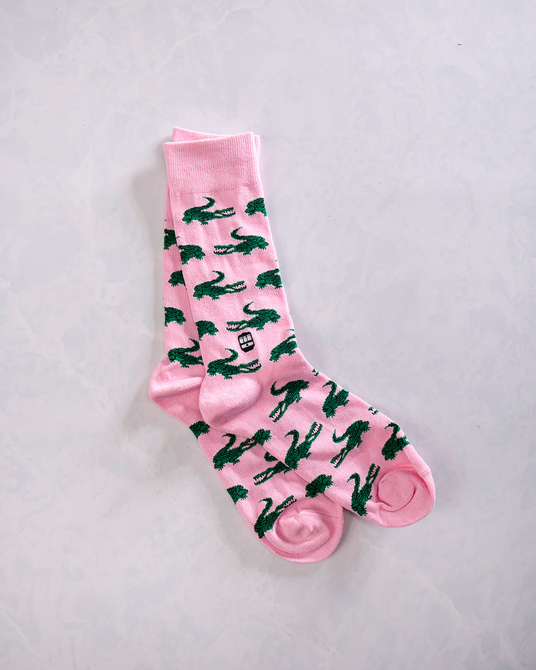 Pair of pink crew socks overlapping on a light background, with a repeating dark green alligator pattern.