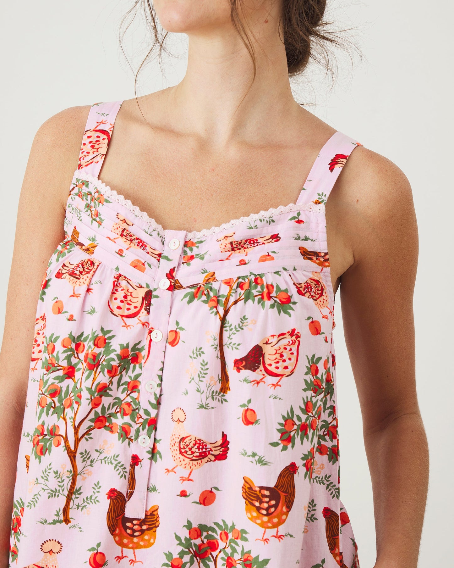 Close-up front view of a ballet-pink nightgown with wide shoulder straps, white buttons, scalloped neckline trim, and a print of hens, fruit, and leafy trees.